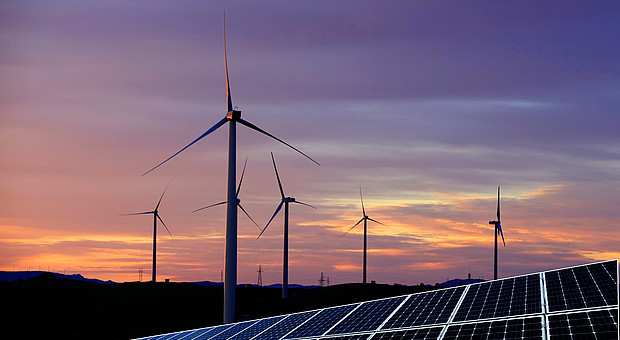 Several wind turbines and an area with solar panels stand in a hilly landscape. In the background, a picturesque sunset with a violet-orange sky.