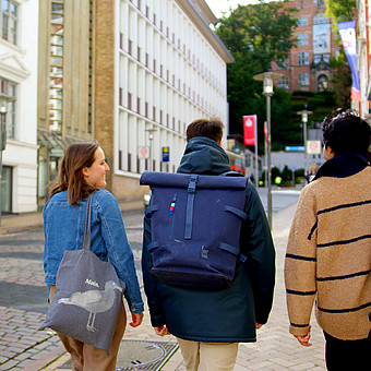 zeigt drei Studierende von Hinten. Sie laufen mit Rucksack die Rathausstraße von Flensburg entlang 