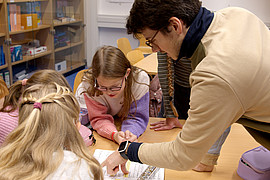 Lehrkraft zeigt einer Gruppe von Kindern an einem Tisch in einer Bibliothek etwas auf einem Blatt Papier.