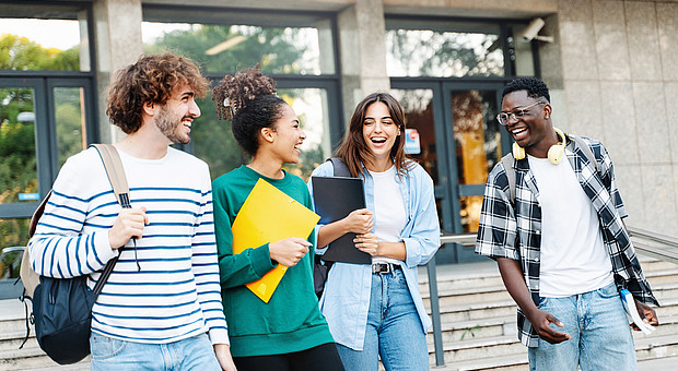 Vier junge Menschen in legerer Kleidung verlassen lachend ein Gebäude. Sie tragen Taschen und Mappen bei sich.