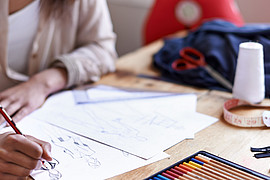 eine junge Designerin zeichnet an einem braunen Schreibtisch an einem Modekonzept mit Bleistift 