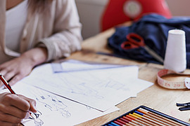 eine junge Designerin zeichnet an einem braunen Schreibtisch an einem Modekonzept mit Bleistift 