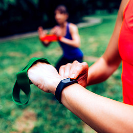 zeigt zwei junge Personen bei Sport Übungen. Person im Vordergrund prüft ihre Smartwatch.
