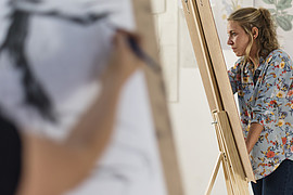 zeigt zwei junge Menschen an einer Staffelei, während eines Kunst Seminars. Der vordere Arm ist leicht verschwommen und skizziert eine Figur.