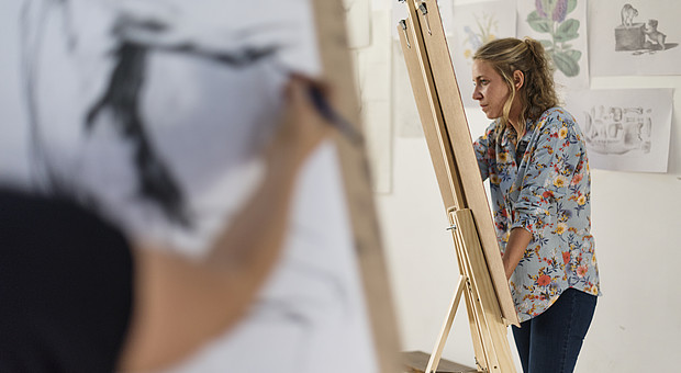 zeigt zwei junge Menschen an einer Staffelei, während eines Kunst Seminars. Der vordere Arm ist leicht verschwommen und skizziert eine Figur.