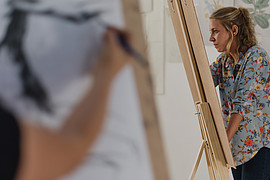 zeigt zwei junge Menschen an einer Staffelei, während eines Kunst Seminars. Der vordere Arm ist leicht verschwommen und skizziert eine Figur.