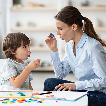 Frau und Kind sitzen an einem Tisch mit bunten Spielsteinen und einem Holzbrett und halten jeweils einen Spielstein in der Hand.