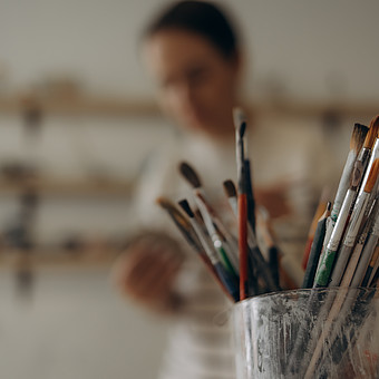 zeigt Pinsel in einem Glas. Im Hintergrund ist verschwommen eine junge Person am Malen. 