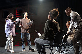 Vier Studierende und eine Dozierende Person befinden sich in einem Proberaumund proben für ein Theaterstück. 