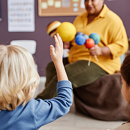 Nahaufnahme einer Rückenansicht von Kindern. Ein kleiner Junge mit erhobener Hand während Lehrkraft erklärt 