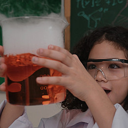 ein Kind mit Laborkleidung hält eine rote Flüssigkeit fasziniert in die Luft. 