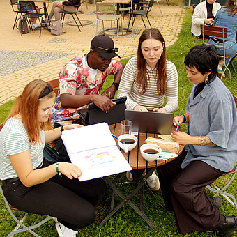 zeigt Studierende draußen auf dem Campus der Europa-Universität Flensburg. Sie trinken Kaffee an einem Tisch. Der Tisch ist versehen mit Laptops und Lernzetteln.  
