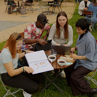 zeigt Studierende draußen auf dem Campus der Europa-Universität Flensburg. Sie trinken Kaffee an einem Tisch. Der Tisch ist versehen mit Laptops und Lernzetteln.  