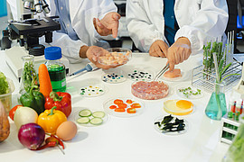 zwei Menschen führen Experimente mit Lebensmitteln durch. Auf einem Tisch stehen unterschiedliche Gemüsesorten 