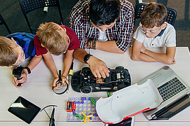 Eine Lehrkraft sitzt mit jungen Schülerinnen und Schülern an einem Tisch und erklärt Technik mit Automodell 