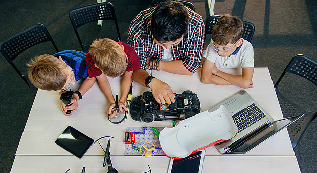 Eine Lehrkraft sitzt mit jungen Schülerinnen und Schülern an einem Tisch und erklärt Technik mit Automodell 