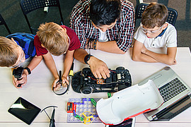 Eine Lehrkraft sitzt mit jungen Schülerinnen und Schülern an einem Tisch und erklärt Technik mit Automodell 