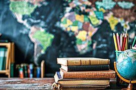 zeigt Bücher und einen Globus auf einem Schreibtisch vor einer blauen Weltkarte im Hintergrund. 