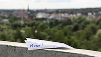 Auf einer Mauer liegt ein Papierflieger mit der Aufschrift "MOIN!". Im Hintergrund ist die Skyline Flensburgs zu sehen.