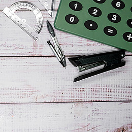 Ein grüner Taschenrechner leigt auf einem Schreibtisch. Darunter ist ein Lineal, Ein Zirkel und ein Tacker zu sehen.  