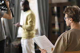Person sitzt auf einem Stuhl und hält ein Blatt Papier, während eine andere Person vor einer Kamera und eine dritte Person im Hintergrund steht.