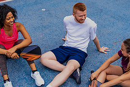 zeigt drei junge Menschen in Sport Kleidung, sie sitzen redend auf einem Sportplatz 