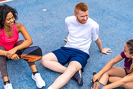 zeigt drei junge Menschen in Sport Kleidung, sie sitzen redend auf einem Sportplatz 