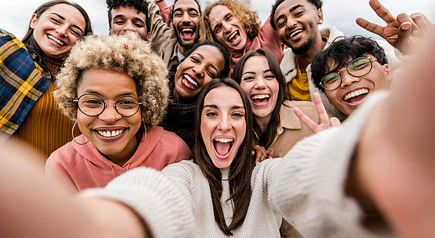 Gruppe junger Menschen steht eng beieinander und macht ein Selfie, einige zeigen Handzeichen wie Peace-Zeichen oder Daumen hoch.