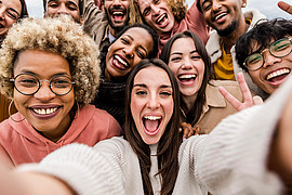 Gruppe junger Menschen steht eng beieinander und macht ein Selfie, einige zeigen Handzeichen wie Peace-Zeichen oder Daumen hoch.