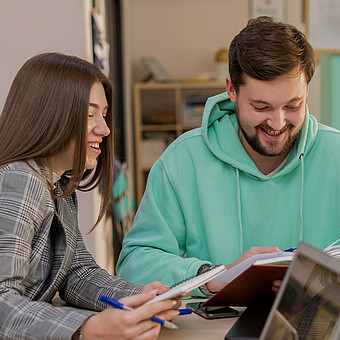  Studierender mit blauem Pulli und Studierende mit Blazer sitzen an einem Tisch mit einem Buch und Notublock  