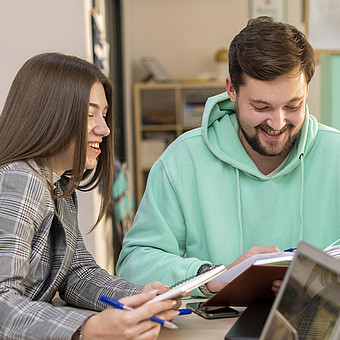  Studierender mit blauem Pulli und Studierende mit Blazer sitzen an einem Tisch mit einem Buch und Notublock  