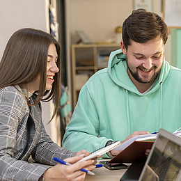  Studierender mit blauem Pulli und Studierende mit Blazer sitzen an einem Tisch mit einem Buch und Notublock  