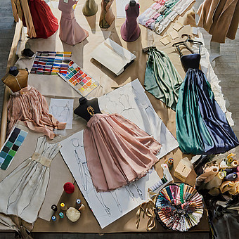 Auf einem Parkett Boden liegen sämtliche Farben an Stoffe, Nähutensilien, Kleider und Skizzierungen zu Kleidungskonzepten. 