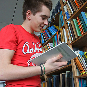 Student steht vor einem Bücherregal und blättert in einem Buch.