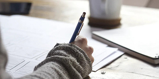 Eine Person schreibt auf einem Blatt Papier, das auf einem Tisch liegt, mit einem Stift.