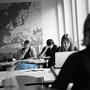 Schwarz/ weiß Foto von Seminarraum, in dem Studierende sitzen. Im Hintergrund steht eine blau akzentuierte Tasche.